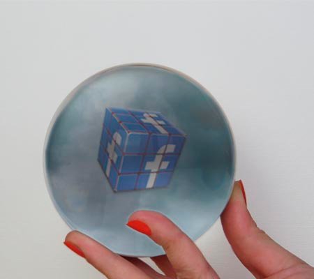 Una mano de mujer sujeta una bola de cristal con nubes oscuras y un cubo de Rubic con el logo de Facebook